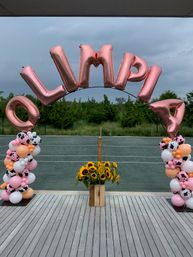 Rose-gold foil letter balloon arch spelling "OLIMPIA" over an outdoor tennis court, flanked by pink, white and cow-print balloon columns and a wooden crate of sunflowers under a cloudy sky.