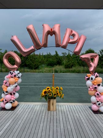 Rose-gold foil letter balloon arch spelling "OLIMPIA" over an outdoor tennis court, flanked by pink, white and cow-print balloon columns and a wooden crate of sunflowers under a cloudy sky.