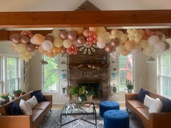 Festive balloon garland in cream, blush and rose-gold arches across a sunlit living room with exposed wood beam, brick fireplace, leather sofas and blue ottomans.