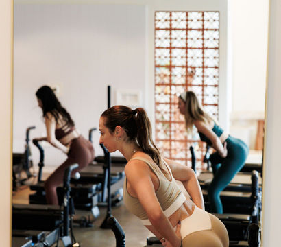 Three women in a bright Pilates reformer class practicing glute and core exercises on reformer machines in a mirrored fitness studio.