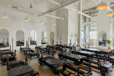 Sunlit modern Pilates reformer studio with rows of black reformer machines, arched mirrors, high ceilings, pendant lights, and concrete floor