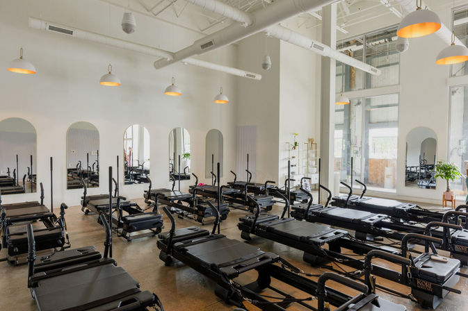 Sunlit modern Pilates reformer studio with rows of black reformer machines, arched mirrors, high ceilings, pendant lights, and concrete floor
