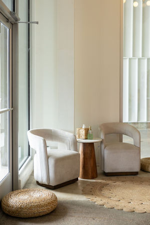 Cozy neutral lounge corner with two curved beige armchairs, small wooden side table, woven jute rugs and pouf by a large window