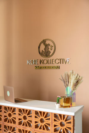 Modern Pilates studio reception with peach walls, gold cactus logo, white countertop over terracotta breeze-block front, laptop, small cactus pot and vase of dried pampas grass.