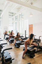 Sunlit modern Pilates studio: women stretching on reformer machines by floor-to-ceiling windows in an airy urban workout space