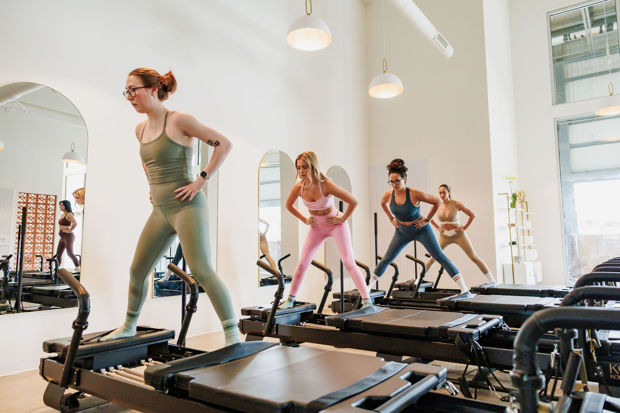 Group reformer Pilates class in a bright modern studio, four women in colorful activewear balancing on reformer machines