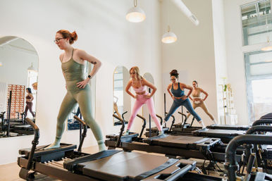 Group reformer Pilates class in a bright modern studio, four women in colorful activewear balancing on reformer machines