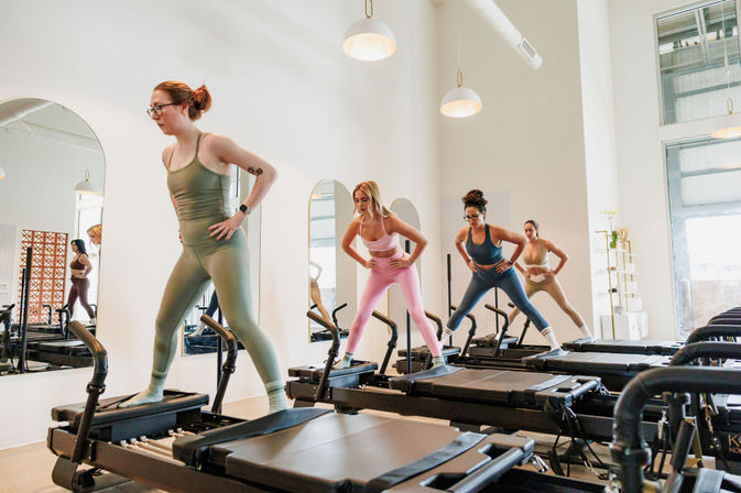 Group reformer Pilates class in a bright modern studio, four women in colorful activewear balancing on reformer machines