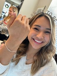 Smiling woman wearing a sparkly "BRIDE TO BE" tiara and white sweater at a home bridal shower, holding up her wrist to show a delicate chain bracelet while a friend is blurred in the living room background.