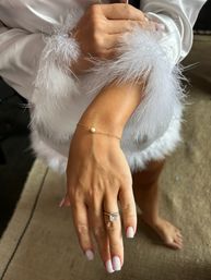 Close-up of a manicured hand in a white satin robe with feather cuffs, showcasing a sparkling diamond engagement ring and a delicate gold chain bracelet with a single pearl — bridal getting-ready detail in an indoor dressing area.