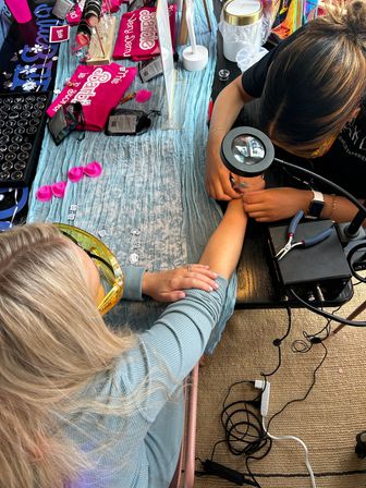 Close-up of an artisan jewelry pop-up: a vendor hand-stamps a custom bracelet on a customer’s wrist under a magnifying lamp at a colorful craft table with charms, pliers, tools and pink promotional shirts.
