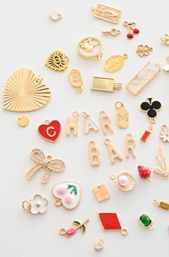 Flat-lay of assorted gold-tone enamel and crystal charm pendants on a white background, including hearts, letters, a bow, cherries, clover, pearl-in-shell, tiny tags and geometric shapes — playful jewelry charms collection.