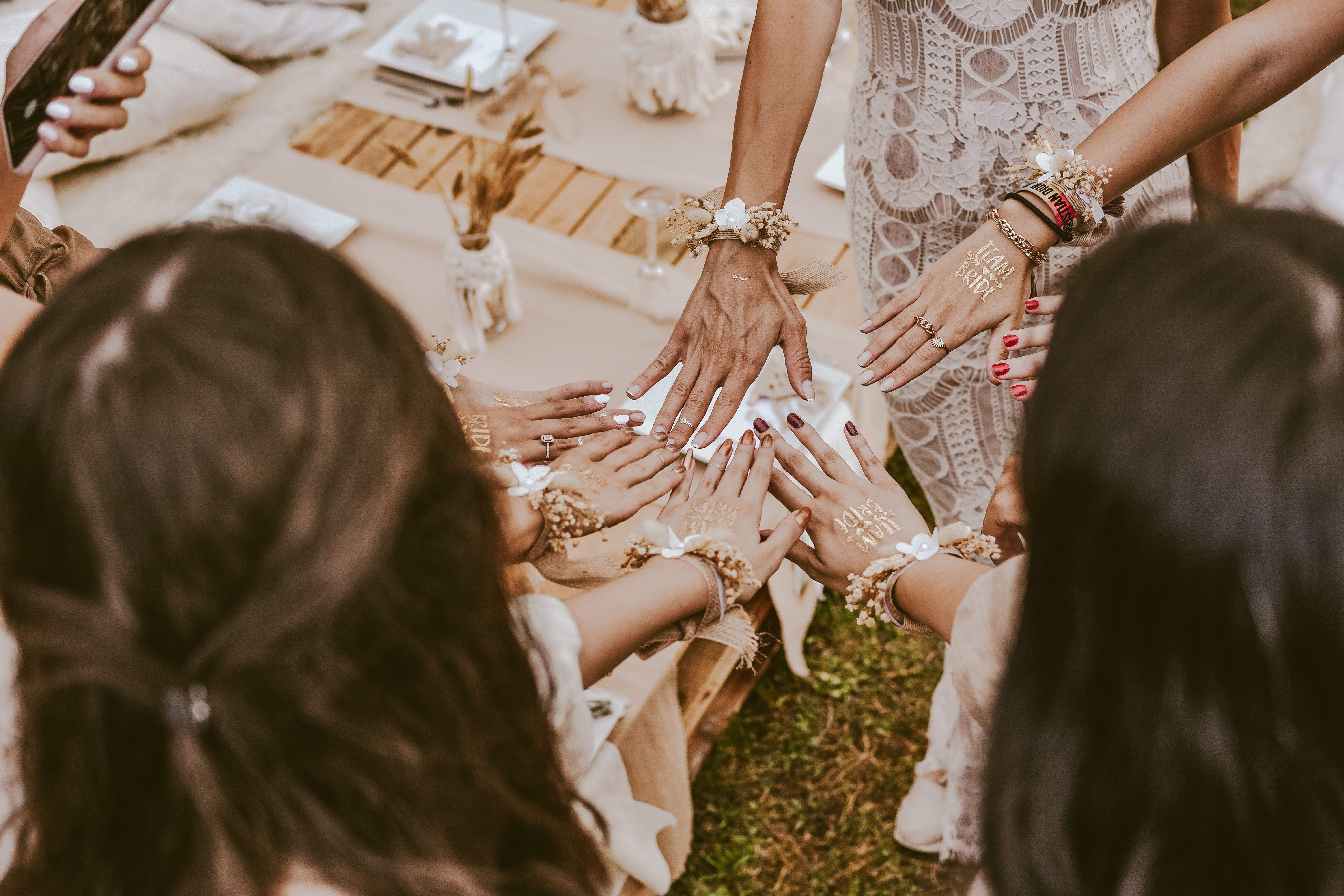 Boho bridal party hands in a circle at an outdoor rustic picnic, bridesmaids and bride wearing lace, floral wrist corsages and gold "Team Bride" temporary tattoos