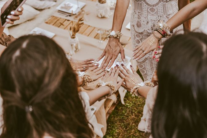 Boho bridal party hands in a circle at an outdoor rustic picnic, bridesmaids and bride wearing lace, floral wrist corsages and gold "Team Bride" temporary tattoos