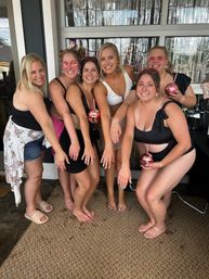 Six friends in swimsuits on a decorated outdoor patio with silver fringe backdrop, holding pink sequin cups and showing rings — fun summer bachelorette vibe
