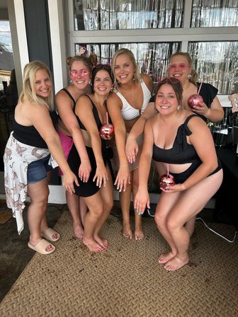 Six friends in swimsuits on a decorated outdoor patio with silver fringe backdrop, holding pink sequin cups and showing rings — fun summer bachelorette vibe