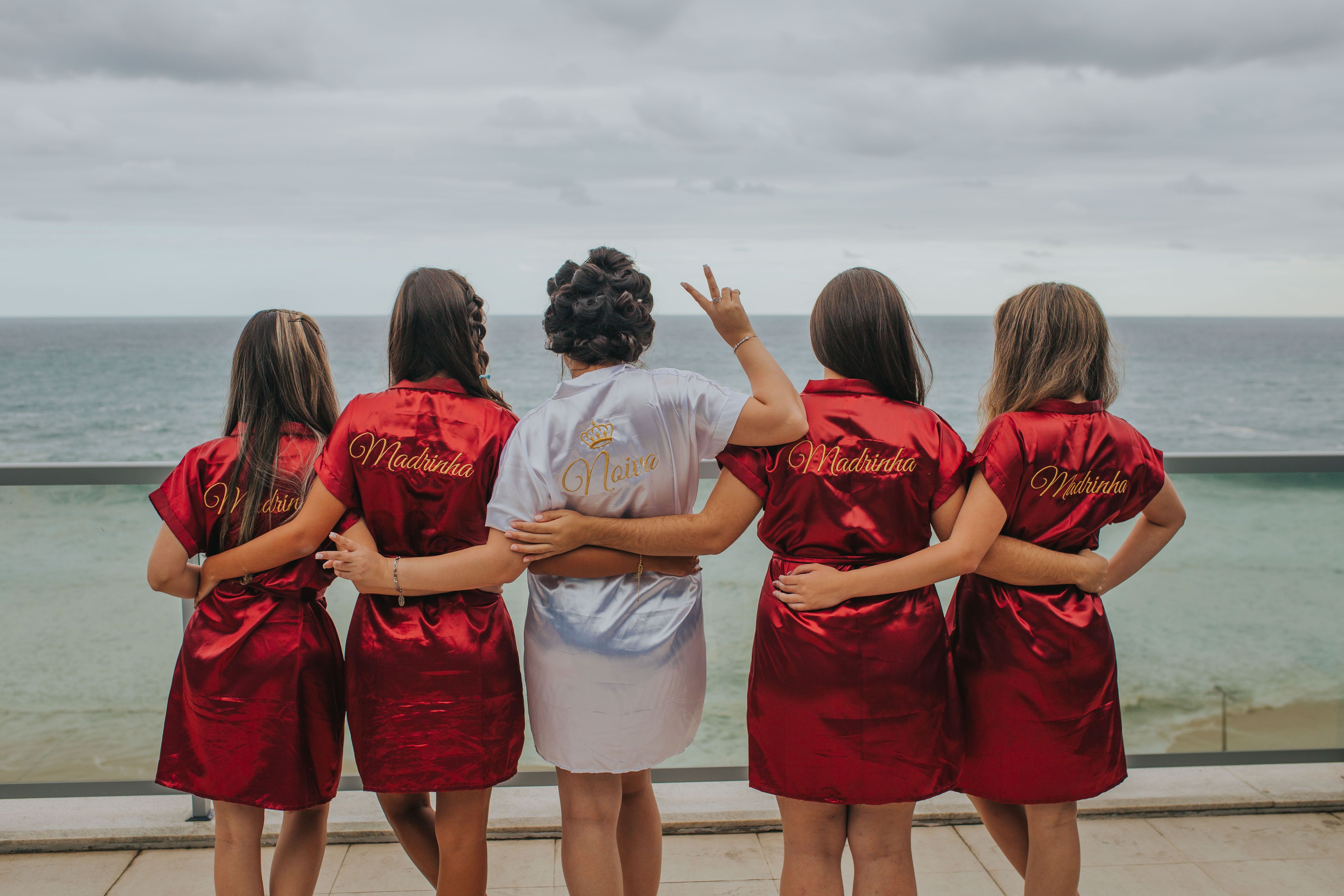 Oceanfront bridal party on a balcony: bride in a white satin robe and four bridesmaids in matching red satin robes with arms around each other facing the sea under a cloudy sky.