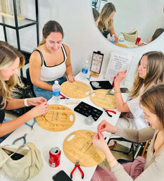 Women at a bright craft studio table in a DIY jewelry-making workshop, arranging beads on wooden bead boards with pliers, trays of beads, water bottles and a round mirror reflection.
