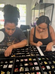 Two women leaning over black compartment jewelry display trays in a bright boutique, carefully selecting sparkly rings, earrings and charms from organized rows.