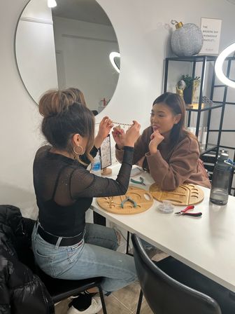 Two women in a modern craft studio inspecting a gold beaded bracelet at a white table surrounded by wooden bead trays, pliers, small beads, a water bottle, round mirror and ring light — casual jewelry-making session.