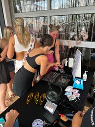 Friends at a bachelorette-party beauty bar on a patio, gathered around a black table with a magnifying lamp, rows of glittery cosmetic samples, yellow safety glasses, wet wipes and a rose-gold fringe backdrop.