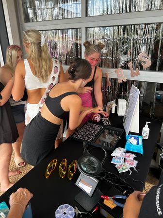 Group of women at an outdoor patio party beauty booth with silver tinsel backdrop, table of cosmetics, yellow protective glasses, magnifying lamp, wipes and small accessories — fun bachelorette-style setup.