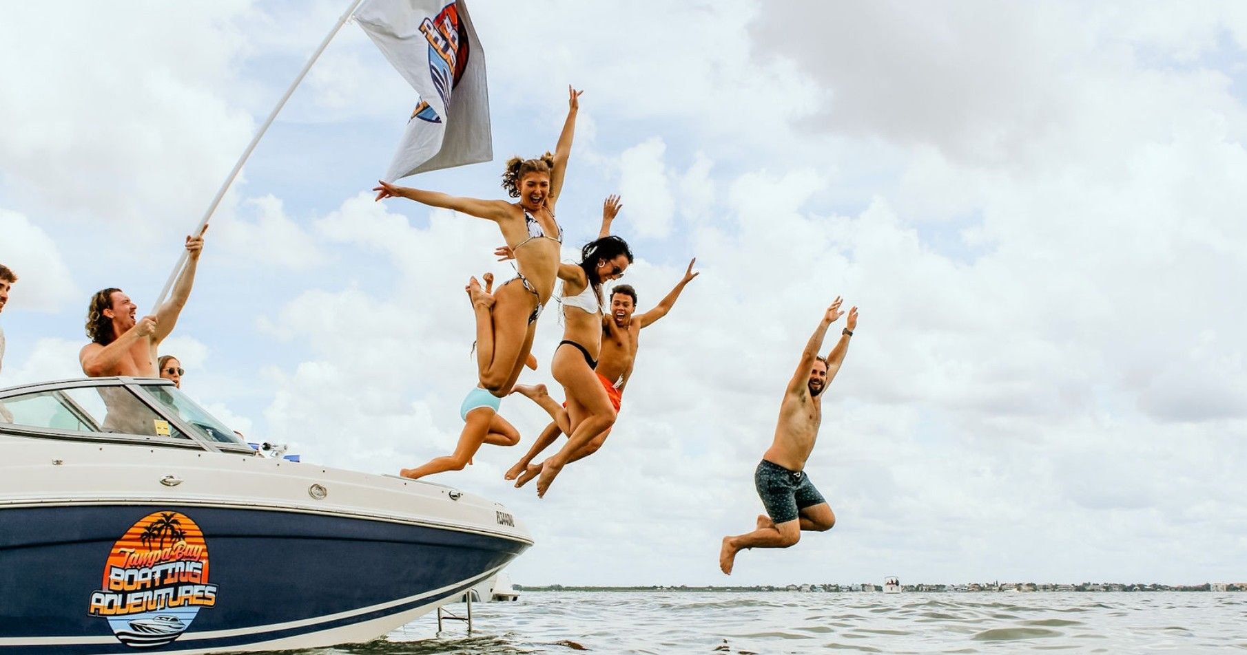 Group of friends jumping off a motorboat into coastal bay waters, laughing in swimwear as a flag waves overhead under a cloudy sky