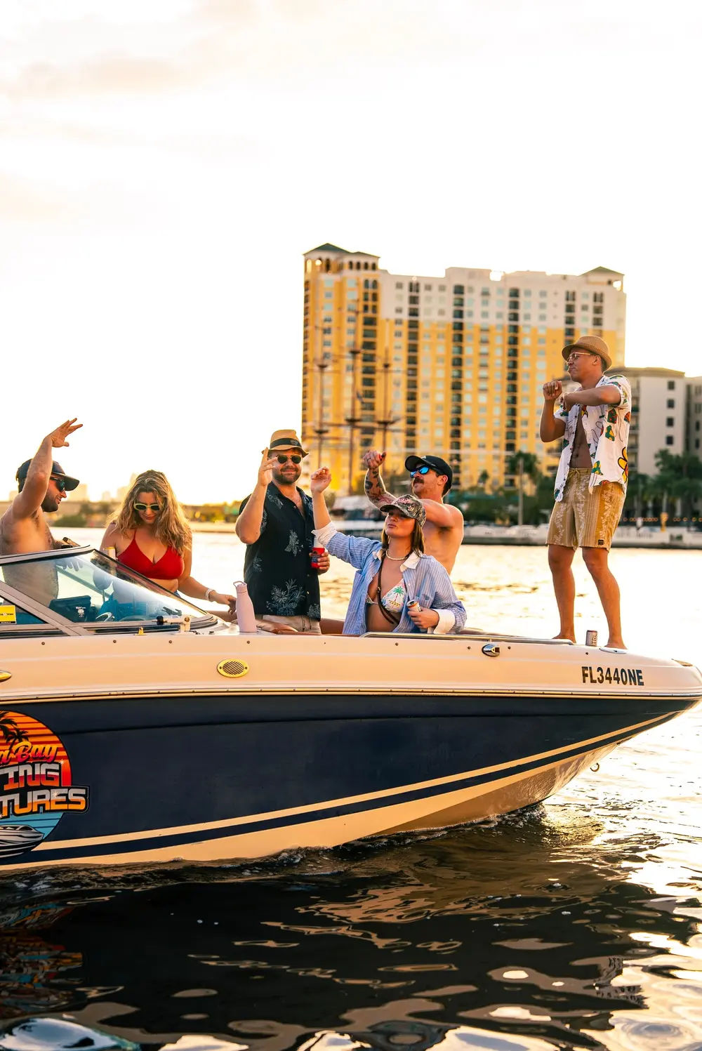 Boat party at sunset on a Florida waterfront — friends in swimwear and sunglasses cheering and dancing on a motorboat with high-rise condos in the background.