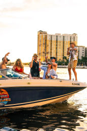 Boat party at sunset on a Florida waterfront — friends in swimwear and sunglasses cheering and dancing on a motorboat with high-rise condos in the background.