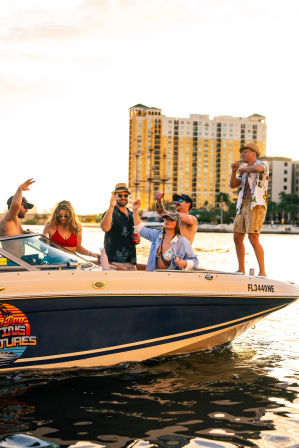 Boat party at sunset on a Florida waterfront — friends in swimwear and sunglasses cheering and dancing on a motorboat with high-rise condos in the background.