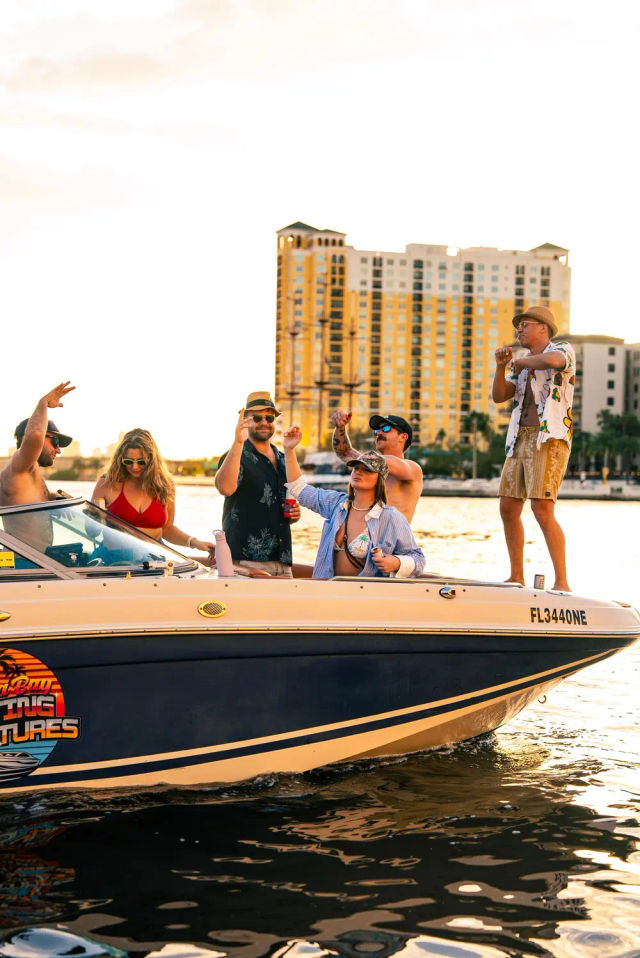Boat party at sunset on a Florida waterfront — friends in swimwear and sunglasses cheering and dancing on a motorboat with high-rise condos in the background.