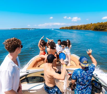 Group of friends partying on a speedboat in turquoise coastal waters with a tropical mangrove shoreline and bright blue summer sky