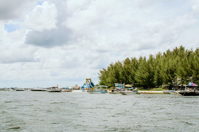 Boats anchored off a sandy beach with a large blue inflatable water slide, colorful umbrellas, pine trees and a cloudy sky.