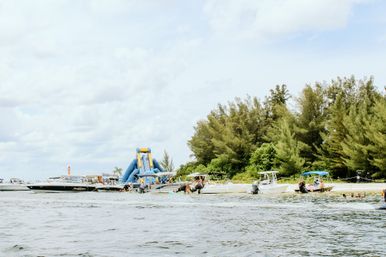 Coastal beach scene with boats anchored off a sandy shore, a large blue inflatable water slide on a floating platform, swimmers in the water, and green trees lining the coastline under a bright cloudy sky.