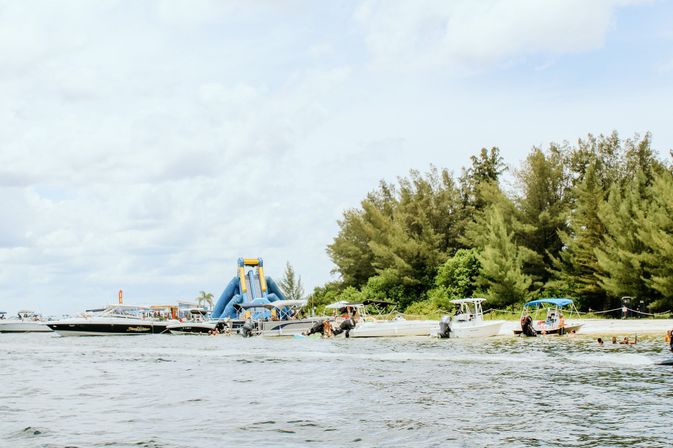 Coastal beach scene with boats anchored off a sandy shore, a large blue inflatable water slide on a floating platform, swimmers in the water, and green trees lining the coastline under a bright cloudy sky.
