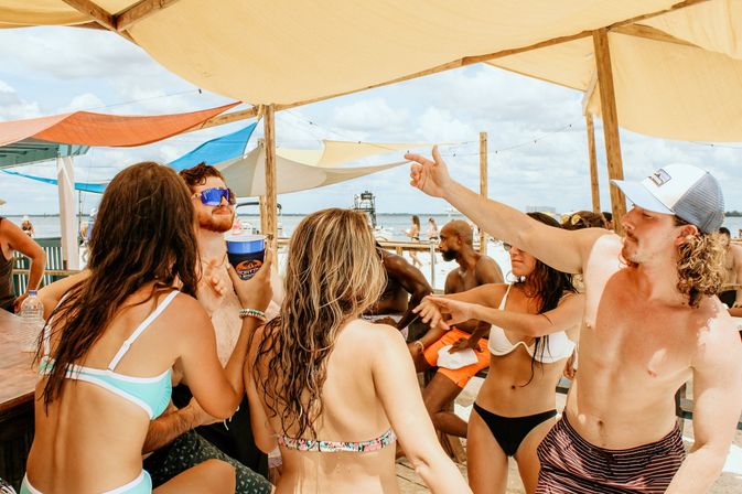 Beachside summer party: friends in swimsuits dancing and holding drinks under colorful sunshade sails on a wooden dock by the ocean with boats and blue sky in the background.