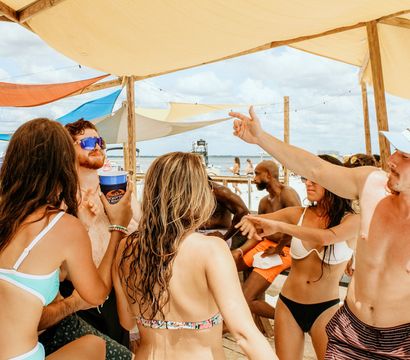 Young adults in swimsuits chatting and holding drinks under colorful shade sails at a sunny beach bar with the ocean and boats in the background.