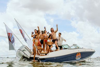 Group of friends at a boat party on a motorboat in shallow coastal waters, people in swimsuits cheering with drinks and waving flags under a blue, cloudy sky