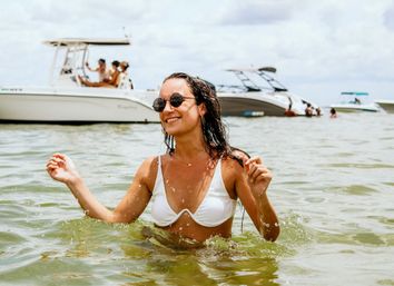Smiling woman in a white bikini and round sunglasses wading in shallow coastal water with several leisure boats in the background on a sunny summer day