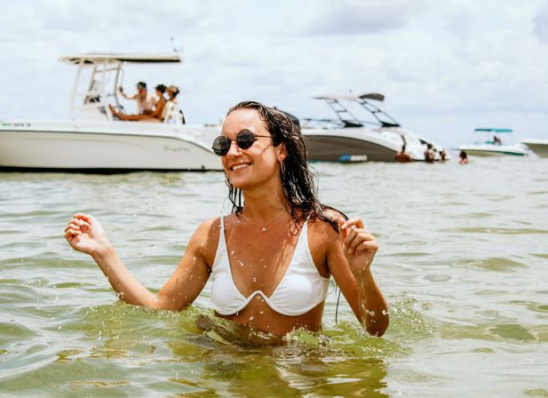 Smiling woman in a white bikini and round sunglasses wading in shallow coastal water with several leisure boats in the background on a sunny summer day