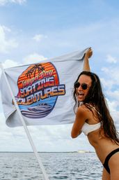 Woman in white bikini and sunglasses laughing while holding a colorful boating flag aboard a boat on a sunny blue bay