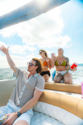 Three friends partying on a sunny motorboat in coastal waters — man in sunglasses holding a can and two women in bikinis (one with tattoos) cheering with drinks against a blue sky and ocean backdrop.