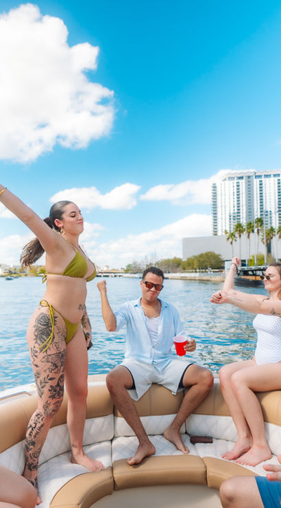Sun-soaked boat party: friends in swimsuits dancing and sipping drinks on a pontoon in a palm-lined urban waterfront with high-rise skyline.
