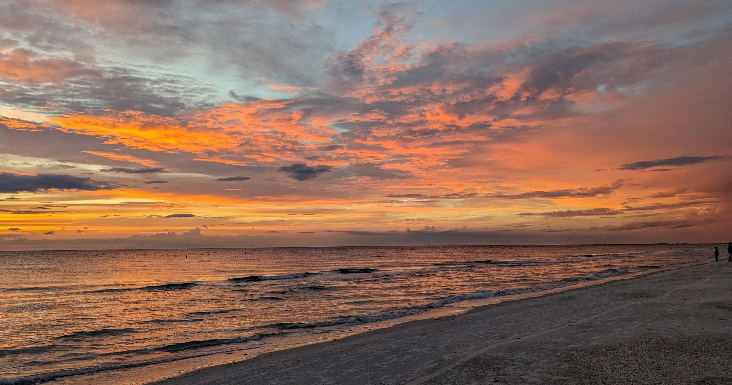 Vibrant orange-pink sunset over a calm ocean with dramatic cloud-filled sky reflecting on gentle waves along a sandy beach and a few distant silhouettes on the shoreline.