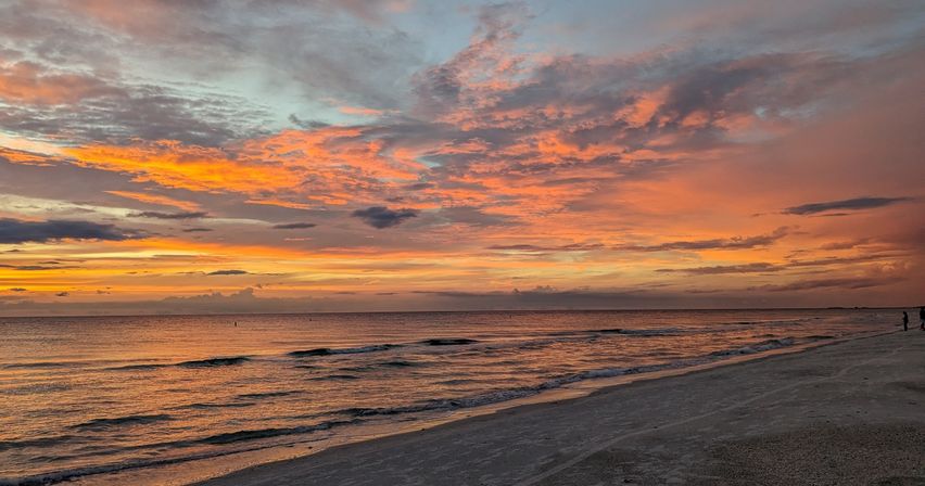 Vibrant orange-pink sunset over a calm ocean with dramatic cloud-filled sky reflecting on gentle waves along a sandy beach and a few distant silhouettes on the shoreline.