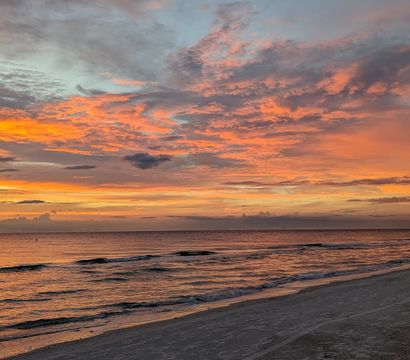 Vibrant orange and pink sunset over a calm ocean and sandy beach, dramatic layered clouds and gentle waves reflecting the colorful sky