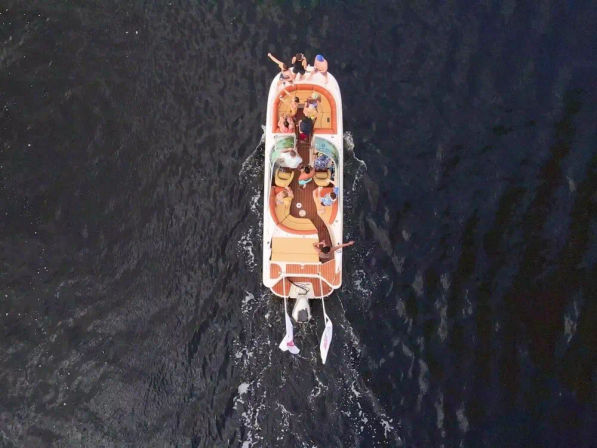 Aerial view of a colorful motorboat with several people relaxing and waving while cruising on dark open water, wake trailing behind