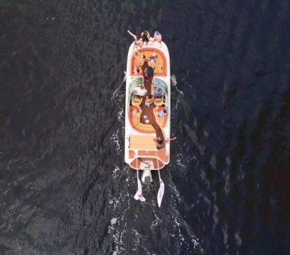Aerial view of a colorful motorboat with several people relaxing and waving while cruising on dark open water, wake trailing behind
