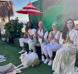 Four people in light clothing seated on a sunny coastal terrace with ocean view, hands in prayer for a seaside meditation session; a white fluffy dog lounges on green turf under a red umbrella and potted plants.