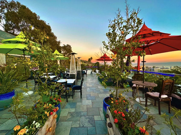 Vibrant sunset rooftop terrace with tiled patio, colorful red and green umbrellas, potted flowers, outdoor dining tables and ocean view.